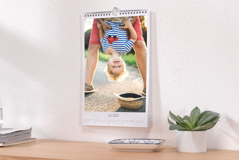 Een vrolijke jongen hangt ondersteboven op een wandkalender in juli 2021. Achter hem zijn benen te zien en op de tafel een kleine plant.