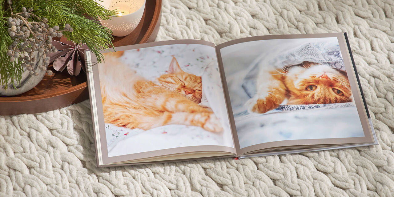 Een CEWE FOTOBOEK in vierkant formaat ligt opengeslagen op een witte deken. Op de dubbele pagina zie je foto's van een rode kat. Links van het fotoboek staat een dienblad met kerstversieringen en kaarsen.