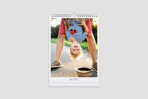 Een kleine jongen hangt vrolijk ondersteboven voor een volwassene die hem vasthoudt, op een zonnig pad. Cewe fotokalender.