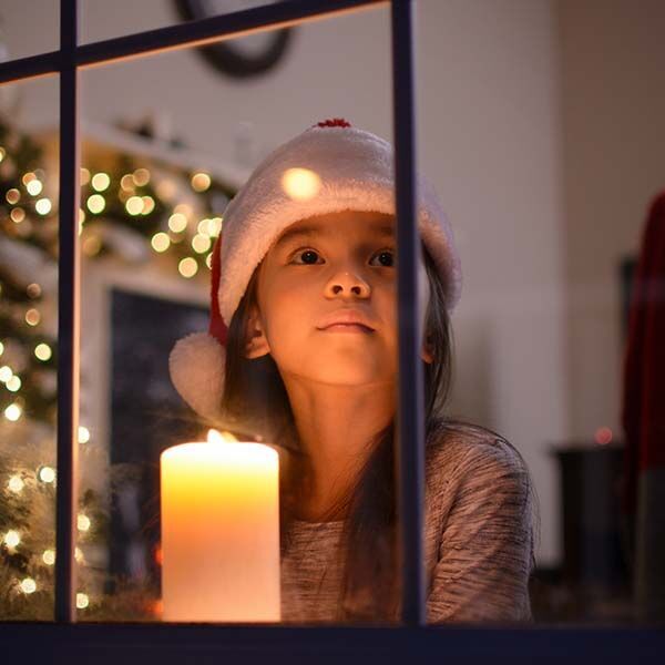 Kind met kerstmuts zit bij het raam bij kaarslicht.